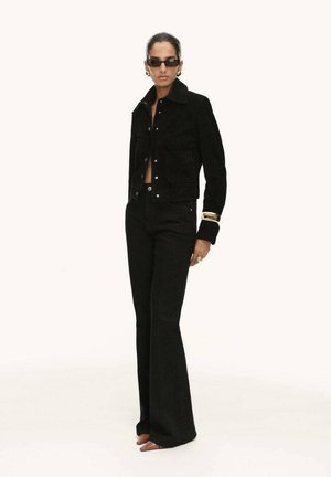 Zwarte corduroy jas met voorzakken, gecombineerd met high-waisted zwarte flared broeken. Model draagt ronde zonnebrillen en gouden accessoires.