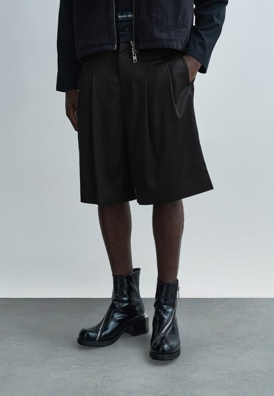 Shorts noirs plissés en tissu texturé, associés à des bottines noires brillantes avec un détail zippé et un talon épais.