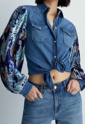 Kvinde iført blå denim cropped skjorte med blomstrede plisserede lange ærmer og matchende blå denimjeans, hænder i forlommerne.