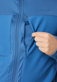 Blaue Jacke mit durchgehendem Reißverschluss, die mit strukturierten oberen Einsätzen und einem hervorgehobenen Logo versehen ist. Die Seitentasche ist teilweise geöffnet und zeigt das Innenfutter sowie den glatten Reißverschluss.