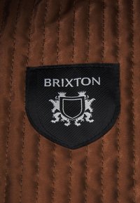 Brixton patch on a brown quilted fabric featuring a black shield design with white lettering and a coat of arms illustration.