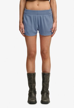 Vrouw draagt lichtblauwe shorts van gestructureerde stof en donkergrijze midkuitlaarzen, staat met ontspannen handen langs haar lichaam.