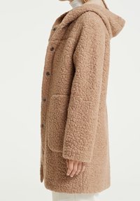 Personne portant un manteau beige texturé mi-long avec de gros boutons, des poches avant et une capuche, vue de côté.