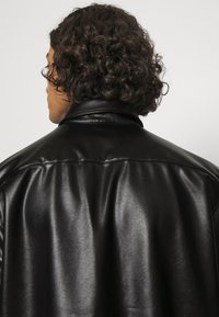 Giacca in pelle nera con una texture liscia e lucida, caratterizzata da un ampio colletto e una vestibilità rilassata, vista da dietro.