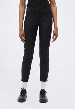 Osoba v priliehavých čiernych nohaviciach, bielych ponožkách a čiernych šnurovacích topánkach, stojaca pred jednoduchým svetlým pozadím.