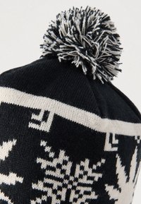 Μαύρο πλεκτό χειμωνιάτικο καπέλο με λευκά σχέδια νιφάδας χιονιού και μαύρο-λευκό πομπόν στην κορυφή, σε λευκό φόντο.
