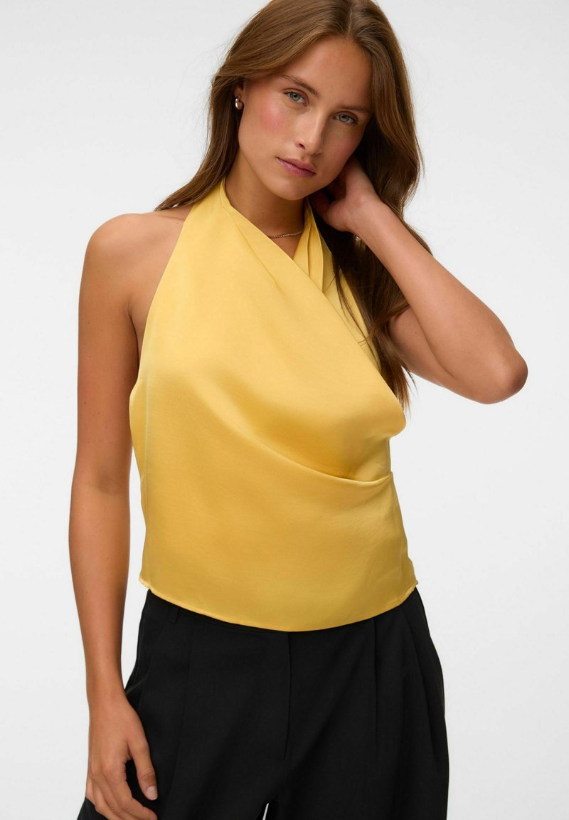 Femme aux longs cheveux bruns portant un top dos-nu jaune et un pantalon noir, posant avec une main touchant son cou sur un fond uni.