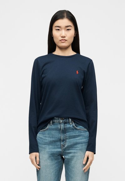 Γυναίκα που φοράει μπλε σκούρο πουκάμισο με μακριά μανίκια και κόκκινο λογότυπο στο στήθος και ανοιχτό μπλε τζιν παντελόνι, στέκεται μπροστά σε ένα απλό λευκό φόντο.