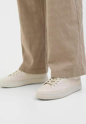 Beige Sneaker aus glattem, strukturiertem Material mit flachen Schnürsenkeln, runder Zehenpartie und einer weißen Gummisohle, kombiniert mit hellbraunen weiten Hosen.