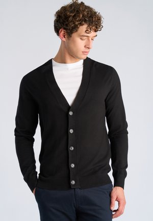 Mann mit lockigem Haar, der eine schwarze, geknöpfte Strickjacke über einem weißen Hemd und dunklen Hosen trägt, schaut nach unten und zur Seite.