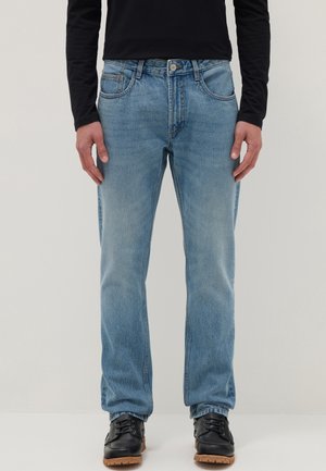 Hombre vistiendo jeans rectos azul claro, camiseta de manga larga negra y zapatos negros con cordones, de pie contra un fondo liso.