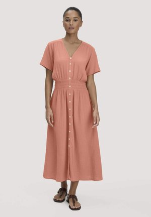 Frau trägt ein lachsrosa Midi-Kleid mit Knöpfen, kurzen Ärmeln und elastischem Bund, kombiniert mit schwarzen Sandalen.