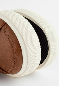 Chaussure en cuir marron avec semelle en caoutchouc blanche texturée et motif de bande de roulement en zigzag noir visible en dessous.