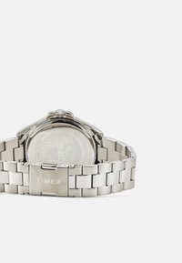 Orologio Timex in acciaio inossidabile argento con bracciale a maglie, cassa rotonda e marchio ben visibile sulla chiusura. Il retro presenta un design a globo.