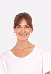 Balta kokvilnas T-krekla ar plašu kaklu. Modelis ar brūniem matiem, nēsā apaļas auskari un smaida uz vienkrāsaina fona.