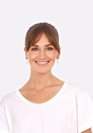 Balta kokvilnas T-krekla ar plašu kaklu. Modelis ar brūniem matiem, nēsā apaļas auskari un smaida uz vienkrāsaina fona.