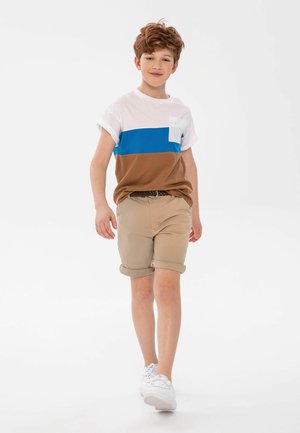 T-shirt à manches courtes bicolore en blanc, bleu et marron, associé à un short beige. Mélange de coton, design simple, poignets retroussés et baskets blanches.