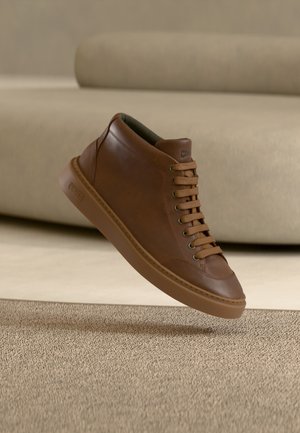 Brown leather sneaker with laces appears to float above a beige carpet, with a soft beige sofa in the blurred background.