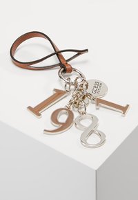 Des boucles en cuir marron fixent les chiffres métalliques "1", "9", "8" et une étiquette circulaire argentée avec l'inscription "GUESS EST.1981" sur une chaîne.