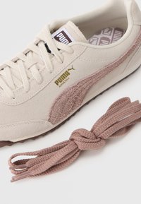 Αθλητικά παπούτσια από σουέτ σε ανοιχτό μπεζ με υφασμένη καφέ ρίγα PUMA. Περιλαμβάνει πλεξούδα ροζ κορδόνι με κόμπο για επιπλέον λεπτομέρεια.