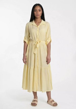 Kobieta stojąca, ubrana w bladożółtą midi sukienkę na guziki z podwiniętymi rękawami i przewiązaną talią, w zestawieniu z beżowymi sandałami z odkrytymi palcami.