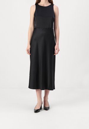 Femme portant une robe midi noire sans manches et des talons en cuir verni noir, debout devant un fond clair uni.
