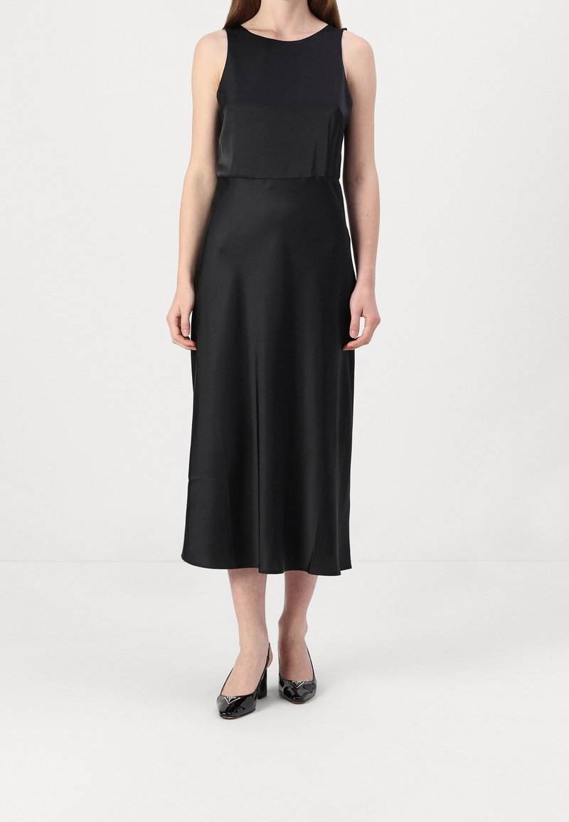 Femme portant une robe midi noire sans manches et des talons en cuir verni noir, debout devant un fond clair uni.