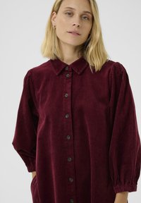 Blonde vrouw in een ruime, lange mouw, bordeauxrode corduroy overhemd met knopen, een kraag en elastische manchetten.
