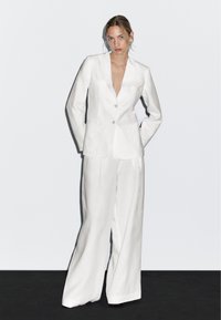 Witte linnen blazer met een geknoopte kraag en twee voorzakken, gecombineerd met een wijde broek, beide van een gestructureerde stof.