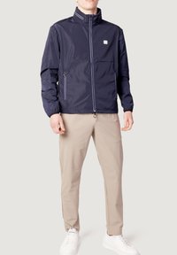 Marineblå vindjakke med høj krave, sidelommer med lynlås, elastiske manchetter og hvide detaljer. Båret over lyse beige bukser og hvide sneakers.