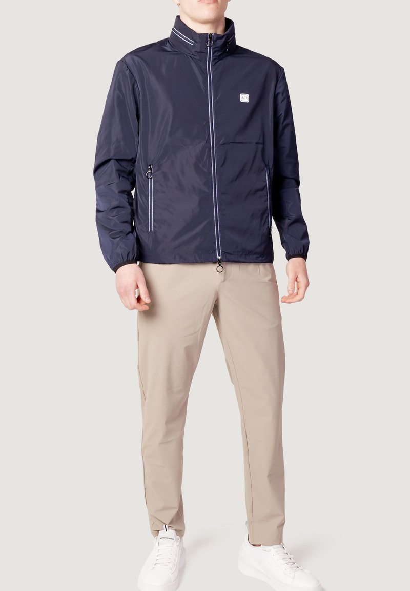 Marineblå vindjakke med høj krave, sidelommer med lynlås, elastiske manchetter og hvide detaljer. Båret over lyse beige bukser og hvide sneakers.