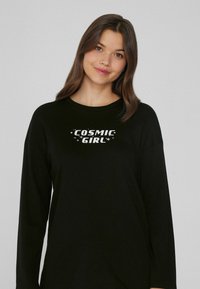 Černé tričko s dlouhým rukávem a kulatým výstřihem, na kterém je nápis "COSMIC GIRL" bílou barvou, obklopený malými hvězdnými akcenty.