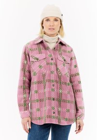 Vrouw in een crèmekleurige beanie, beige coltrui, roze patroon knoop fleece jack en blauwe spijkerbroek, staand tegen een witte achtergrond.