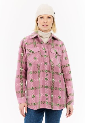 Vrouw in een crèmekleurige beanie, beige coltrui, roze patroon knoop fleece jack en blauwe spijkerbroek, staand tegen een witte achtergrond.