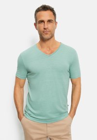Lichtgroene V-hals t-shirt van een zachte, gestructureerde stof met korte mouwen. Model staat met de handen in de zakken en draagt een beige broek.