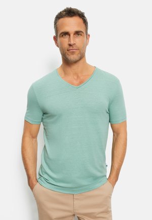 T-shirt verde chiaro a V in un morbido tessuto testurizzato con maniche corte. Il modello sta in piedi con le mani in tasca, indossando pantaloni beigi.
