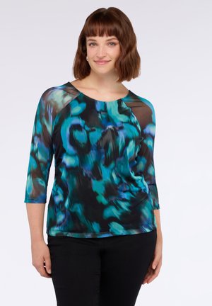 Donna con capelli castano di media lunghezza che indossa una blusa a maniche tre quarti blu e nera con motivo astratto e pantaloni neri.