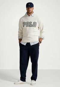 Hvit genser med «POLO» i fete svarte bokstaver, overlappet en blå og hvit skjorte med krage. Marineblå bukser og hvite joggesko fullfører antrekket.