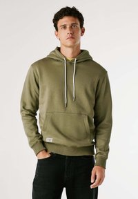 Olivengroene hoodie van een zachte stof, met een voorzak, koordjes en geribde manchetten. Zwarte jeans maken de look compleet.