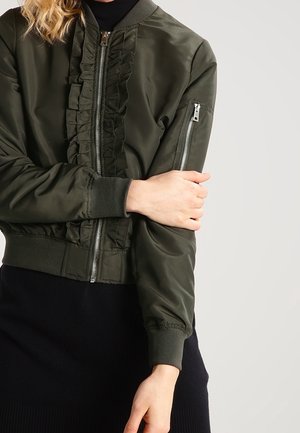 Nainen, jolla on päällään tummanvihreä bomber-takki, jossa on röyhelöyksityiskohta etuosan vetoketjun varrella ja vasemmassa hihassa vetoketjullinen tasku.