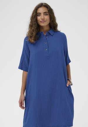 Jonge vrouw met golvend haar, gekleed in een loszittende, blauwe jurk met kraag en knopen, staand met één hand in haar zak tegen een effen achtergrond.