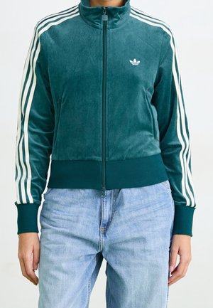 Persoon draagt een teal zip-up Adidas-jas met witte strepen op de mouwen en lichtblauwe jeans, staat met handen ontspannen naast het lichaam.