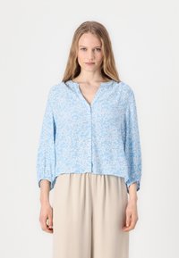 Blusa button-up a maniche lunghe di colore azzurro chiaro, con scollatura a V, caratterizzata da un motivo floreale bianco e polsini arricciati, abbinata a pantaloni beige.
