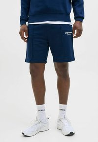 Bermudas deportivas en azul marino, de mezcla de algodón, con un bolsillo lateral y branding en blanco. Combinadas con zapatillas blancas y calcetines acanalados.