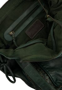 Interieur van een groene canvas tas met een leren label, een ritsvak en oogjesdetails langs de rand. Stevige constructie zichtbaar.