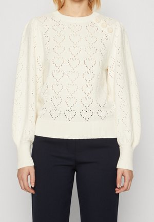 Pull en maille blanc avec motif œillet en forme de cœur, trois boutons décoratifs sur l'épaule gauche, manches longues, porté avec un pantalon noir.