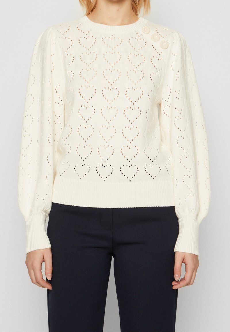 Pull en maille blanc avec motif œillet en forme de cœur, trois boutons décoratifs sur l'épaule gauche, manches longues, porté avec un pantalon noir.