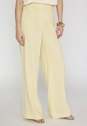 Femme portant un pantalon taille haute et jambes larges de couleur jaune clair, des sandales beige à bouts ouverts, un bracelet en or et des bagues aux deux mains.