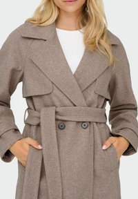 Beige trenchcoat i uldblanding med store revers, dobbeltknaplukning og bælte i livet; har sidelommer og struktureret stof.