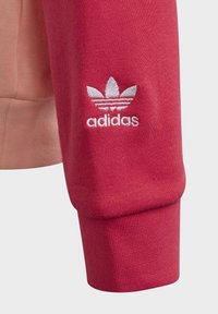Red sweatshirt with textured fabric, ribbed cuffs, and an embroidered white Adidas logo featuring three leaf design on the sleeve.